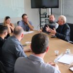 Na imagem aparecem três homens de costas para a câmera, o prefeito e uma vereadora de Piracicaba, o presidente da Associação de Médicos-veterinários da região, e o presidente do CRMV-SP. O prefeito apoio a mão no rosto e faz anotações em um papel branco enquanto presta atenção à fala e gestual do presidente do Conselho. Estão todos sentados ao entorno de uma mesa retangular de madeira.