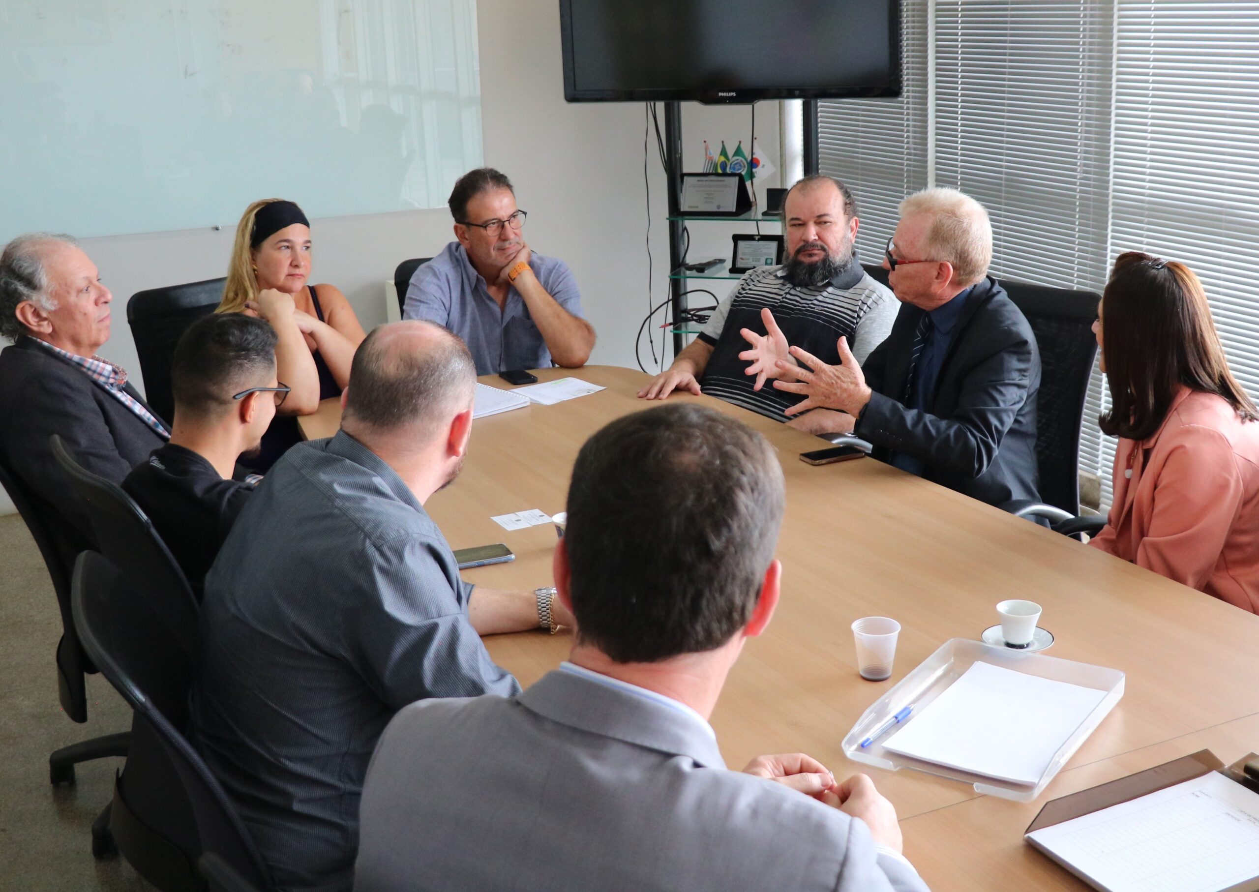 Na imagem aparecem três homens de costas para a câmera, o prefeito e uma vereadora de Piracicaba, o presidente da Associação de Médicos-veterinários da região, e o presidente do CRMV-SP. O prefeito apoio a mão no rosto e faz anotações em um papel branco enquanto presta atenção à fala e gestual do presidente do Conselho. Estão todos sentados ao entorno de uma mesa retangular de madeira.
