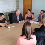 Na foto aparecem seis homens e duas mulheres sentados ao entorno de uma mesa retangular de madeira. Na cabeceira o prefeito de Piracicaba, que apoia a mão no rosto e faz anotações em um papel branco. Quem fala é o integrante da Comissão de Saúde Ambiental e médico-veterinário da região. Ele se dirige ao prefeito. Os demais observam.