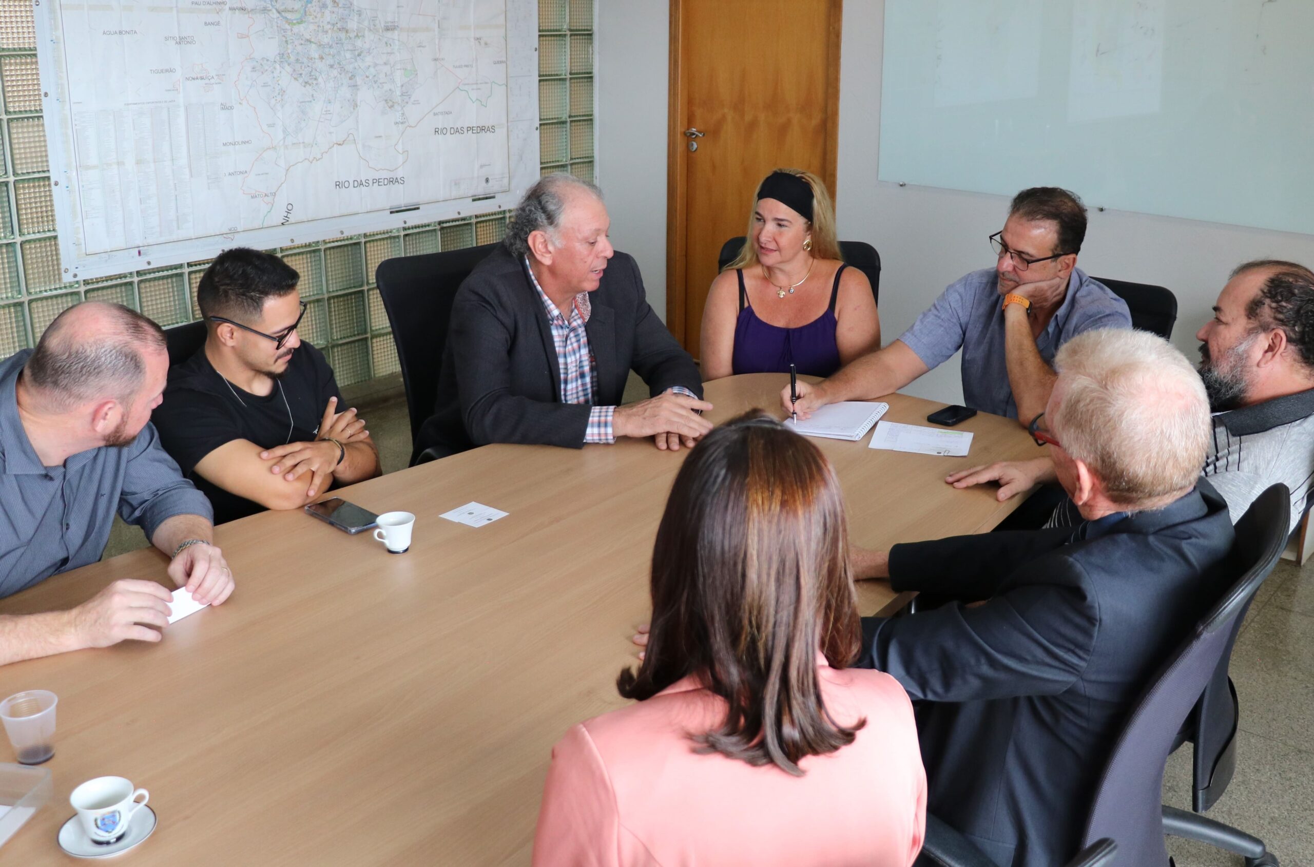 Na foto aparecem seis homens e duas mulheres sentados ao entorno de uma mesa retangular de madeira. Na cabeceira o prefeito de Piracicaba, que apoia a mão no rosto e faz anotações em um papel branco. Quem fala é o integrante da Comissão de Saúde Ambiental e médico-veterinário da região. Ele se dirige ao prefeito. Os demais observam.