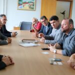 Na imagem aparecem representantes do CRMV-SP e da Associação de Médicos-veterinários de Piracicaba conversando com o secretário de Saúde municipal e uma vereadora. Estão sentados no entorno de uma mesa retangular de madeira. Ao fundo há um quadro na parede com a pintura de uma casa.