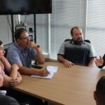 A foto traz cinco homens e uma mulher sentados ao entorno de uma mesa retangular de madeira. dois deles estão de costas para a câmera. O presidente do CRMV-SP fala e gesticula para o prefeito de Piracicaba, que está com a mão apoiada no rosto e faz anotações em um papel branco.