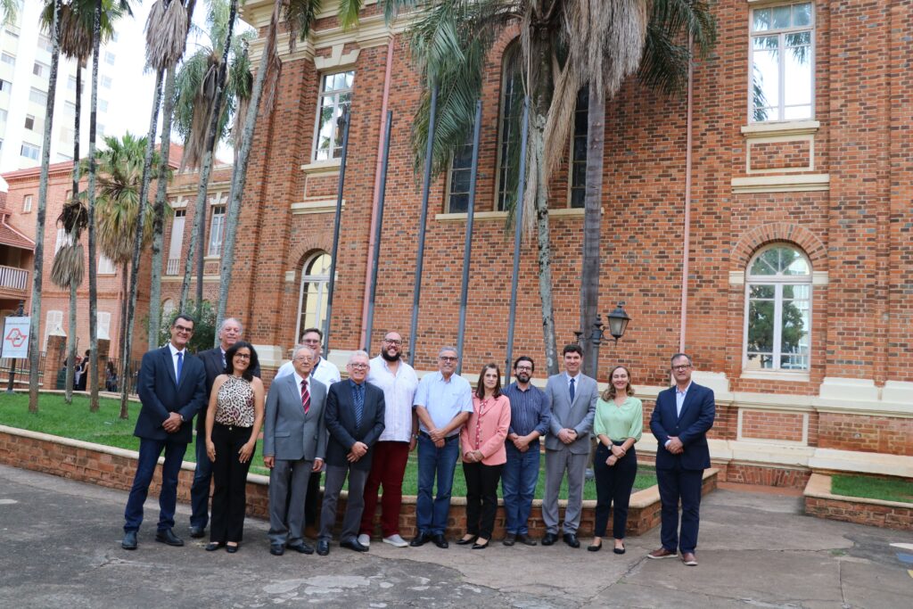 Na foto aparecem os representantes do CRMV-SP e da Unimep. Estão todos de pé, posam e sorriem para a câmera, e estão posicionados em frente a uma construção história com tijolos de barro aparentes cercada por uma jardim com palmeiras. São 10 homens e três mulheres no total.