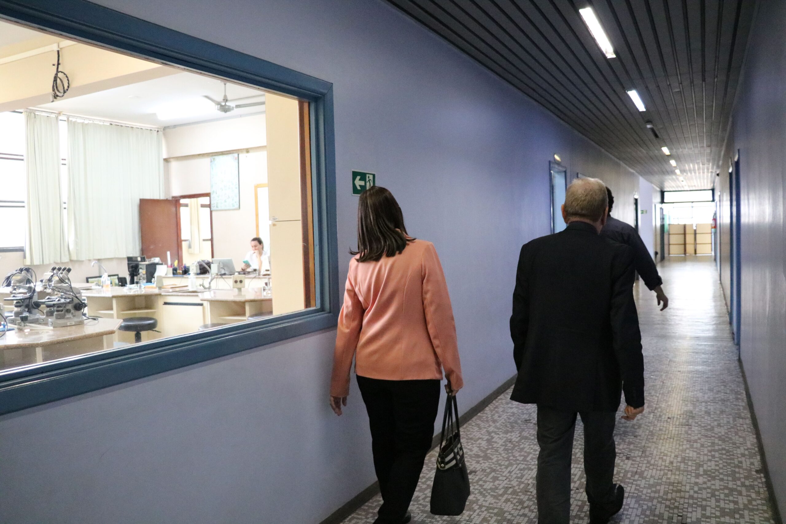 Na foto aparece a tesoureira e o presidente do CRMV-SP de costas em um corredor de salas de aula da Unimep. Na parede a esquerda há uma grande janela de vidro em que é possível observar um dos laboratórios da instituição.