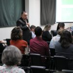 Na imagem aparecem profissionais sentados em cadeiras, de costas para a câmera. Em pé, o integrante da Comissão Técnica de Saúde Pública Veterinária, Mário Ramos. Ele está vestindo termo cinza e uma gravata com listradas vermelha e dourada. Ele está ao microfone.