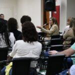 Na imagem aparece em meio a plateia, a integrante da Comissão Técnica de Educação do Conselho, Kátia Saraiva. Ela está sentada, gesticula e segura um microfone. A profissional veste um casaco bege e está de perfil.