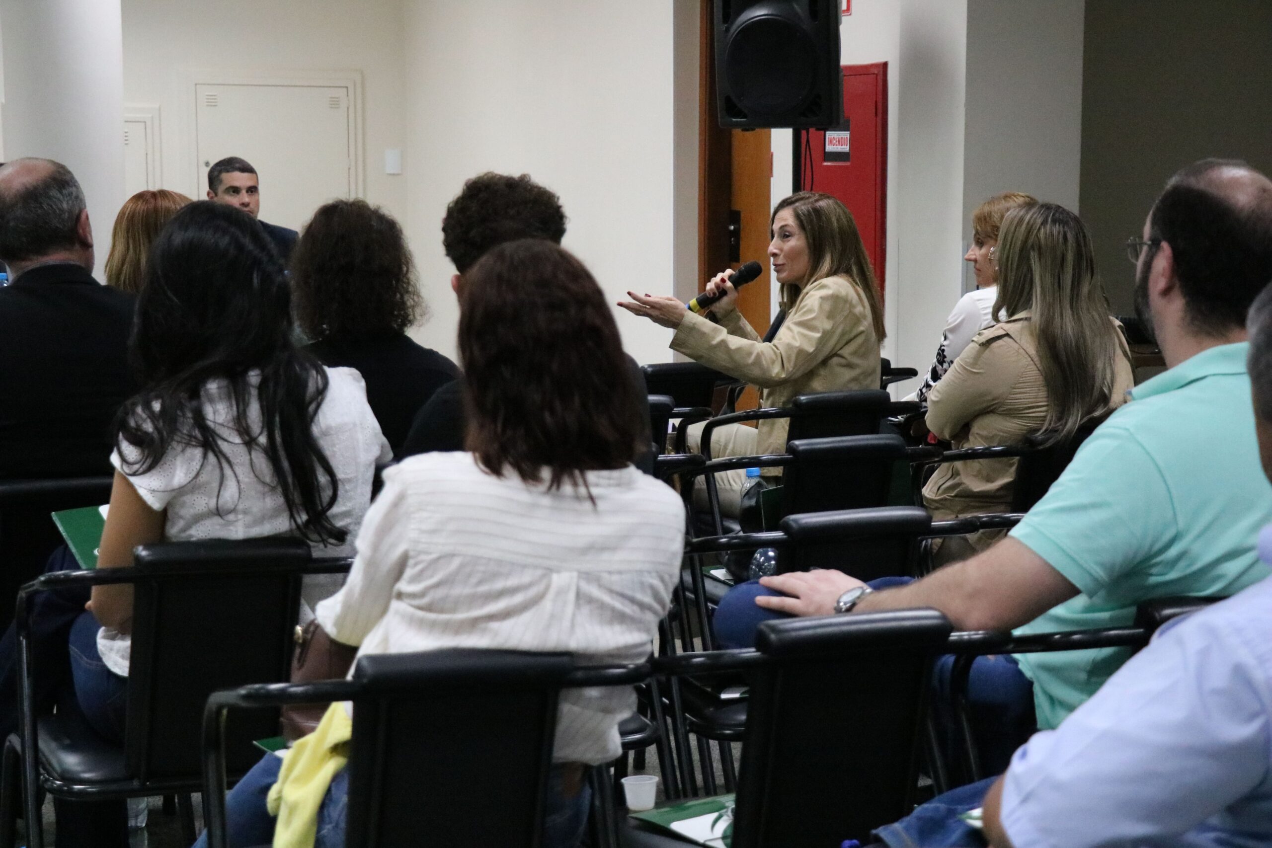 Na imagem aparece em meio a plateia, a integrante da Comissão Técnica de Educação do Conselho, Kátia Saraiva. Ela está sentada, gesticula e segura um microfone. A profissional veste um casaco bege e está de perfil.