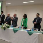 Na foto aparece a uma mesa com toalhas brancas com detalhes verdes e arranjo de flores. Atrás dela esta a diretoria do CRMV-SP, o assessor jurídico e administrativo, e o diretor técnico. Ao microfone, o presidente do Conselho dá as boas-vindas a todos.