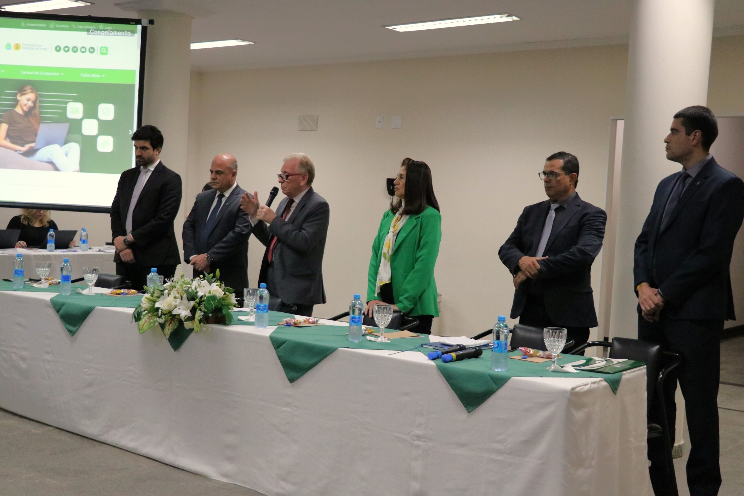 Na foto aparece a uma mesa com toalhas brancas com detalhes verdes e arranjo de flores. Atrás dela esta a diretoria do CRMV-SP, o assessor jurídico e administrativo, e o diretor técnico. Ao microfone, o presidente do Conselho dá as boas-vindas a todos.