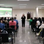 Na foto aparece um auditório lotado, com profissionais sentados e voltados à frente, onde está a chefe do setor de Eventos do CRMV-SP, dando início ao cerimonial de boas-vindas do evento. Ela é loira, de pele clara, usa camisa e calça pretas e sapato nas cores preta e branca. Na foto aparece ao fundo também uma mesa com toalha branca com um arranjo de flores e um telão em que é projetada a campanha de recadastramento.