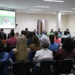 Na imagem aparece um auditório lotado de profissionais. Eles estão de costas para a câmera, sentados em cadeiras e dispostos de frente para a diretoria do Conselho, que está a frente sentada atrás de uma mesa com toalha branca e arranjo de flores. Na sala há ainda um telão em que se destaca o site do CRMV-SP com banner sobre a campanha de recadastramento.