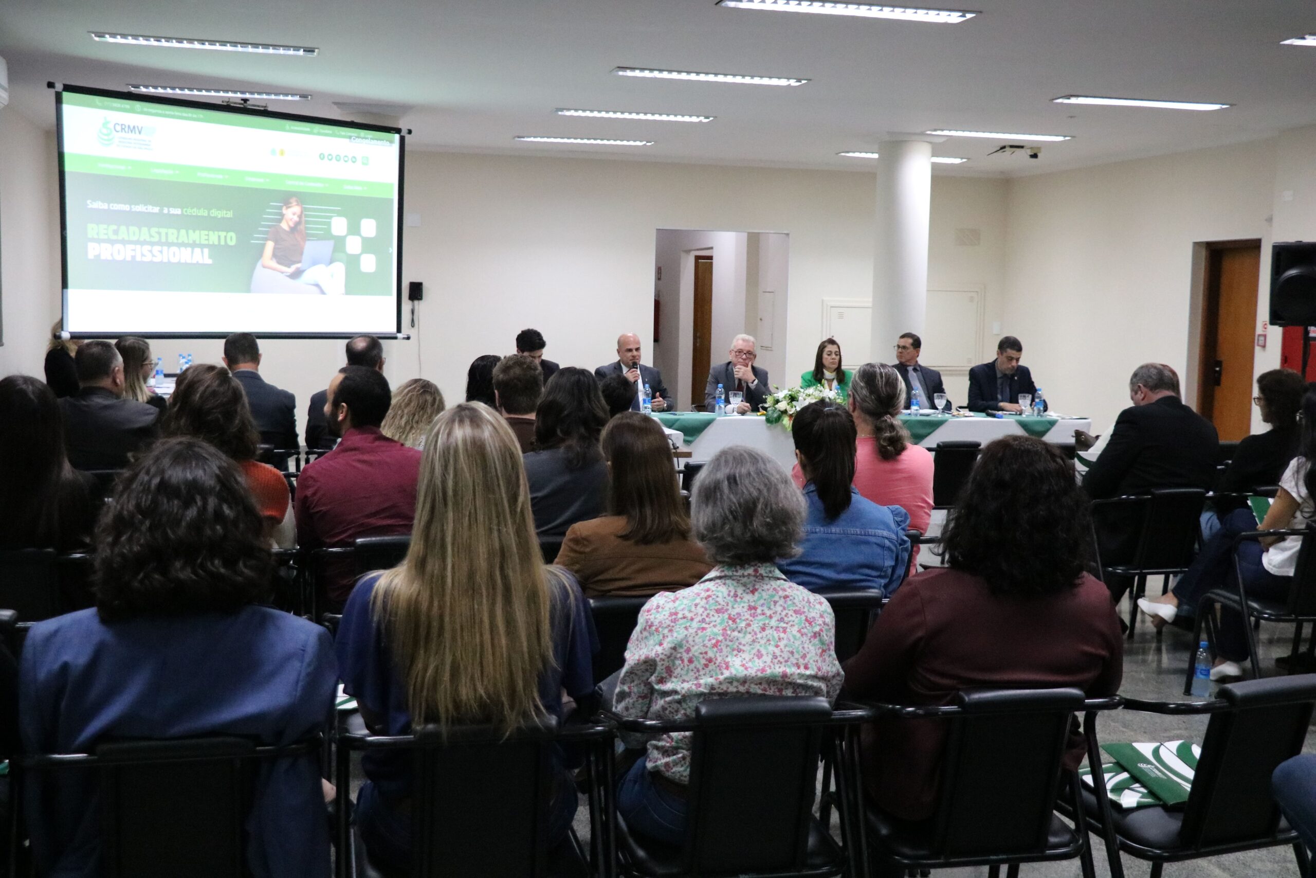 Na imagem aparece um auditório lotado de profissionais. Eles estão de costas para a câmera, sentados em cadeiras e dispostos de frente para a diretoria do Conselho, que está a frente sentada atrás de uma mesa com toalha branca e arranjo de flores. Na sala há ainda um telão em que se destaca o site do CRMV-SP com banner sobre a campanha de recadastramento.