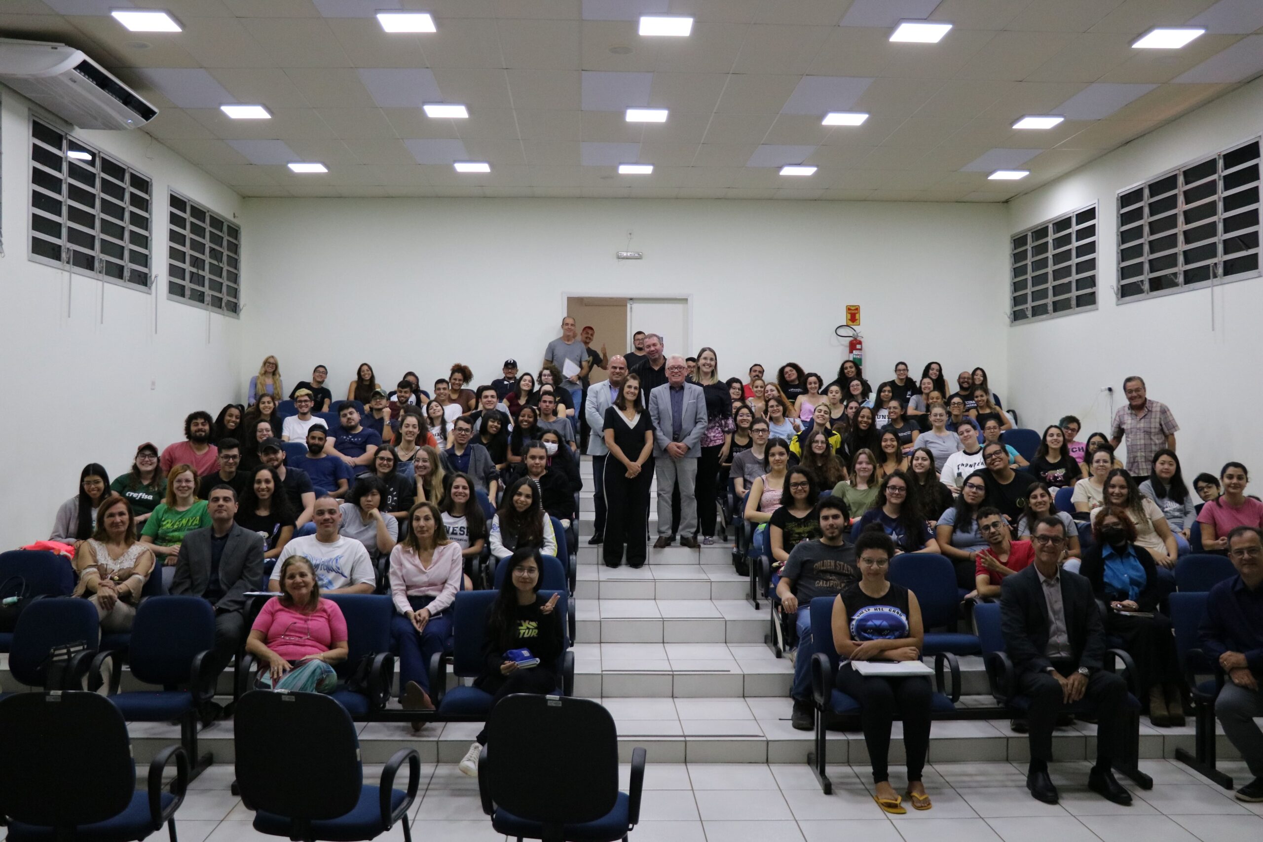 Mais de 120 estudantes da graduação em Medicina Veterinária da Unesp estão sentados nas cadeiras do auditório do Hospital Veterinário. No corredor central estão de pé o presidente, o vice-presidente e a tesoureira do CRMV-SP, acompanhados da representante regional da autarquia em Araçatuba e do representante da Feveresp. Todos posam e sorriem para a foto.