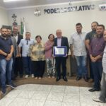 Na imagem o presidente do CRMV-SP aparece segurando a placa com o título de cidadão benevolente ao lado de sua mãe e vereadores da cidade de Sebastianópolis do Sul. Atrás deles os brasões da república e do município com os dizeres Poder Legislativo entre aplicado na parede do plenário.