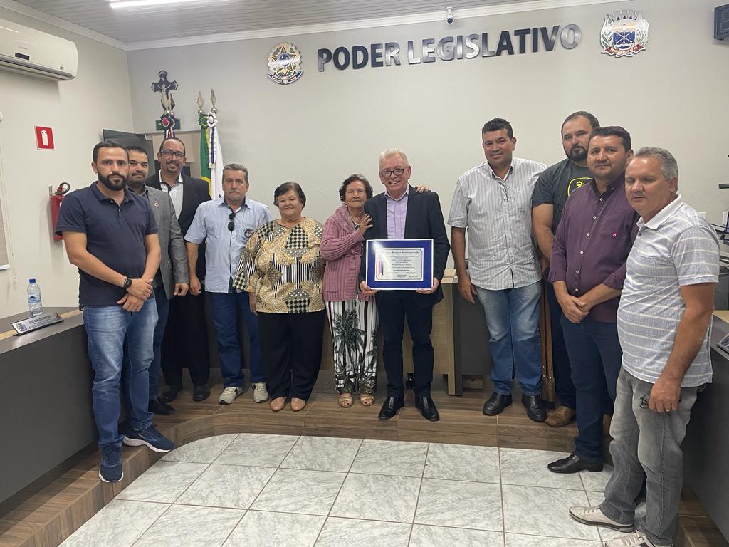 Na imagem o presidente do CRMV-SP aparece segurando a placa com o título de cidadão benevolente ao lado de sua mãe e vereadores da cidade de Sebastianópolis do Sul. Atrás deles os brasões da república e do município com os dizeres Poder Legislativo entre aplicado na parede do plenário.