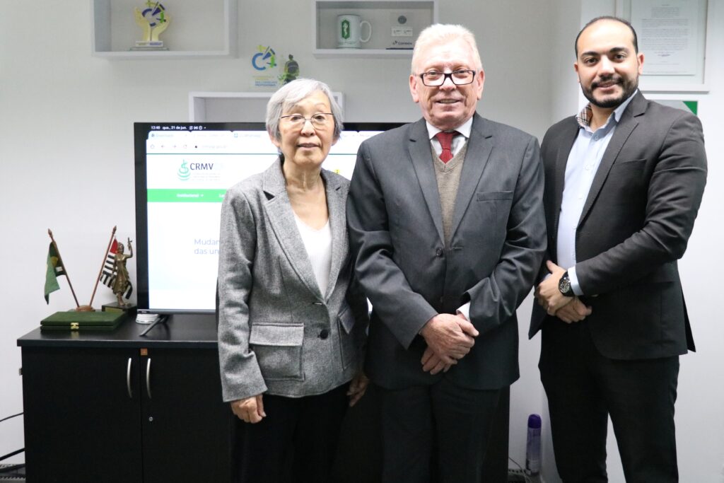 Na foto há dois homens e uma mulher. Eles posam para a foto, olhando e sorrindo para a câmera. Estão em frente a uma televisão em que aparece o site do CRMV-SP, com a sua logomarca.