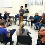 Na foto há 14 pessoas sentadas em carteiras escolares formando uma roda de conversa. Em pé está o secretário de Bem-estar Animal de Mogi Guaçu. O secretário é um homem jovem, moreno, de cabelos curtos e óculos de aro preto.