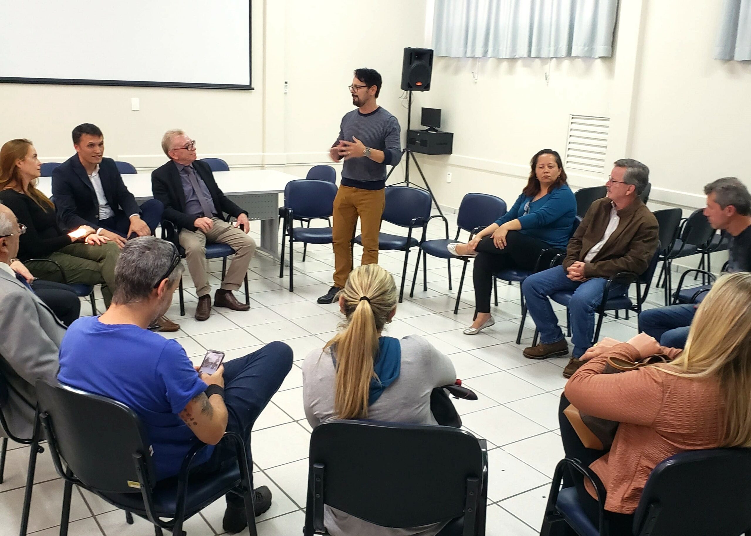Na foto há 14 pessoas sentadas em carteiras escolares formando uma roda de conversa. Em pé está o secretário de Bem-estar Animal de Mogi Guaçu. O secretário é um homem jovem, moreno, de cabelos curtos e óculos de aro preto.