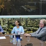 Na imagem aparecem apenas o prefeito e a vice-prefeita de Araçatuba em conversa com o presidente e a tesoureira do CRMV-SP. Eles estão sentados ao entorno de uma mesa marrom e em frente a um painel com fotos de vista aérea da cidade. A vice-prefeita está com um belo sorriso.