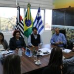 Na imagem aparecem de costas para a câmera a representante regional, a tesoureira e o presidente do CRMV-SP, eles estão sentados ao entorno de uma mesa retangular marrom. Do outro lado, voltados para a câmera e atentos a fala do presidente do Conselho, estão integrantes da Secretaria de Saúde de Araçatuba, o secretário de Meio Ambiente, o prefeito e a vice-prefeita. Na sala lá um painel com fotos aéreas da cidade, uma janela grande em que aparecem árvores e as bandeiras do estado de São Paulo, do Brasil e de Araçatuba.