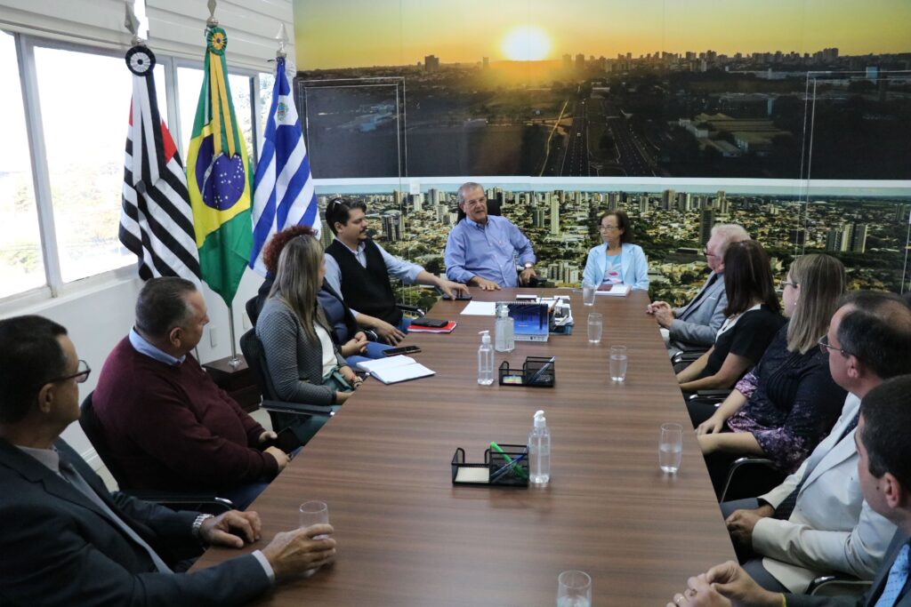 Representantes do CRMV-SP estão reunidos ao entorno de uma grande mesa marrom na Prefeitura de Araçatuba. O prefeito e a vice-prefeita estão na cabeceira da mesa. Ao fundo um painel com imagem de um por do sol e vista aérea da cidade. Há na sala também as bandeiras do estado de São Paulo, do Brasil e do município de Araçatuba. Estão todos sentados e atentos a fala do prefeito, um senhor grisalho, vestindo camisa azul e utilizando óculos.