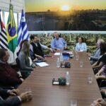 Representantes do CRMV-SP estão reunidos ao entorno de uma grande mesa marrom na Prefeitura de Araçatuba. O prefeito e a vice-prefeita estão na cabeceira da mesa. Ao fundo um painel com imagem de um por do sol e vista aérea da cidade. Há na sala também as bandeiras do estado de São Paulo, do Brasil e do município de Araçatuba. Estão todos sentados e atentos a fala do prefeito, um senhor grisalho, vestindo camisa azul e utilizando óculos.
