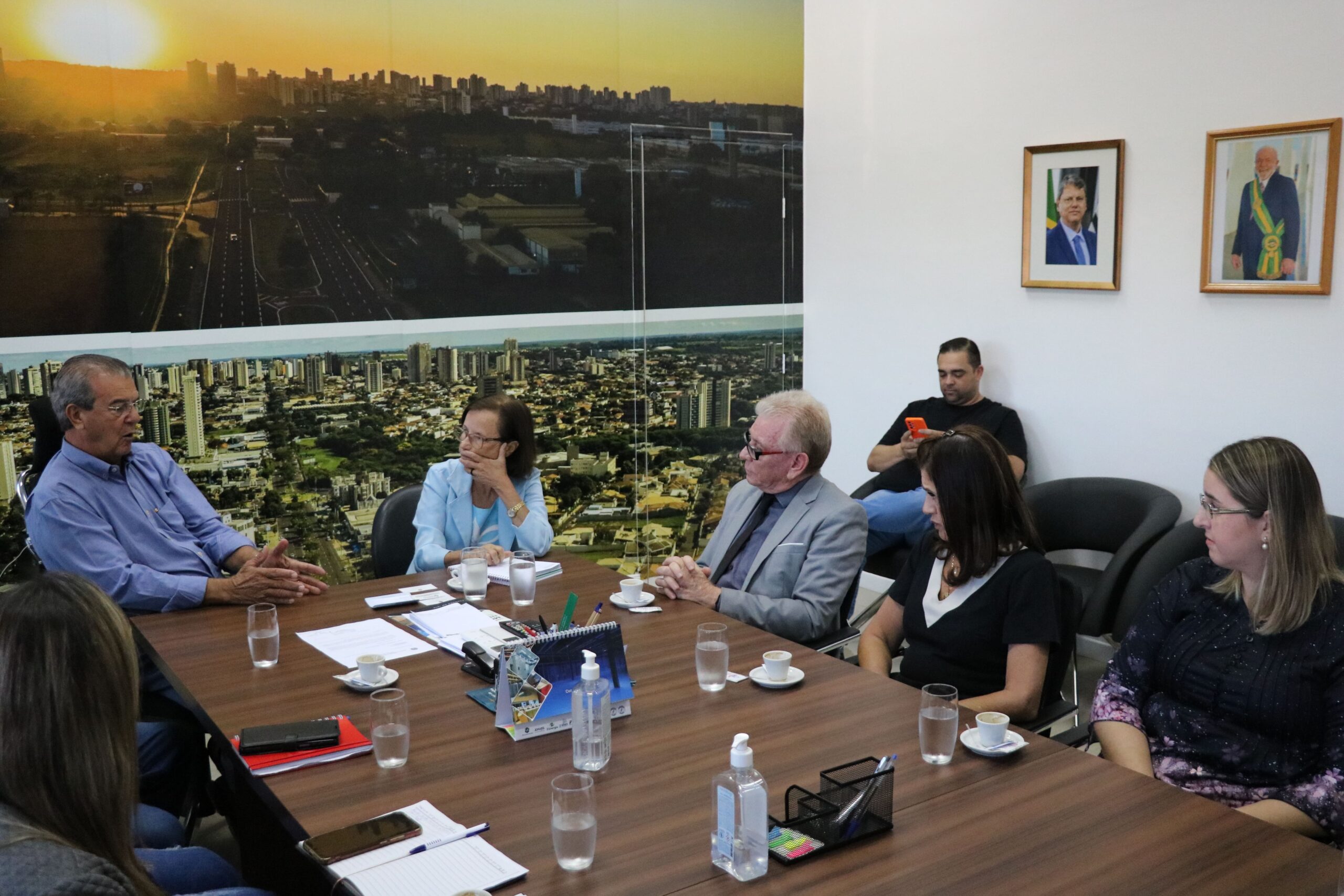 A imagem mostra a sala de reunião do Gabinete do prefeito de Araçatuba em outro ângulo. Estão na foto o prefeito e a vice-prefeita da cidade, bem como o presidente, a tesoureira e a representante regional do CRMV-SP. Eles estão sentados ao entorno de uma mesa retangular marrom e atentos a fala do prefeito. Em uma parede da sala há um painel com fotos aéreas da cidade, e em outra, dois quadros com fotos do governado do Estado, e do presidente do País.