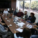 Nesta imagem, aparecem 13 pessoas sentadas ao entorno de uma mesa retangular marrom. São representantes da prefeitura de Araçatuba e do CRMV-SP. Por este ângulo, na cabeceira está o secretário de governo do município. Ao fundo, uma televisão grande desligada, a imagem de "Nossa Senhora Aparecida", e uma janela ampla em que é possível ver árvores e alguns poucos carros.