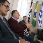 Na foto aparecem três homens, sendo que o do meio está gesticulando enquanto fala, e ao fundo também há uma mulher sentada em um sofá preto. Há ainda as bandeiras do estado de São Paulo, do Brasil e de Araçatuba.