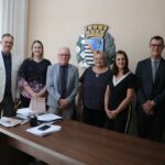 Na imagem há oito pessoas, sendo três mulheres. A foto registra o encontro da comitiva do CRMV-SP com a presidente da Câmera de Vereadores. Eles sorriem e posam lado a lado atrás de uma mesa de madeira e em frente ao brasão da cidade.