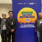 Na imagem há três pessoas, entre elas apenas uma mulher. Eles posam ao lado de um painel da 17ª Conferência Nacional de Saúde, com os dizeres "Amanhã vai ser outro dia!". Eles são médicos-veterinários do CFMV e do CRMV-SP.