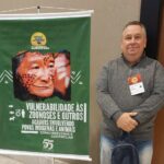 Na imagem aparece o integrante da Comissão Técnica de Saúde Pública do CRMV-SP. Ele é um homem de meia idade, com cabelos curtos e grisalhos. Ao lado dele há um banner verde sobre zoonoses e saúde indígena. No material há a imagem de um indígena com o rosto pintado tradicionalmente.