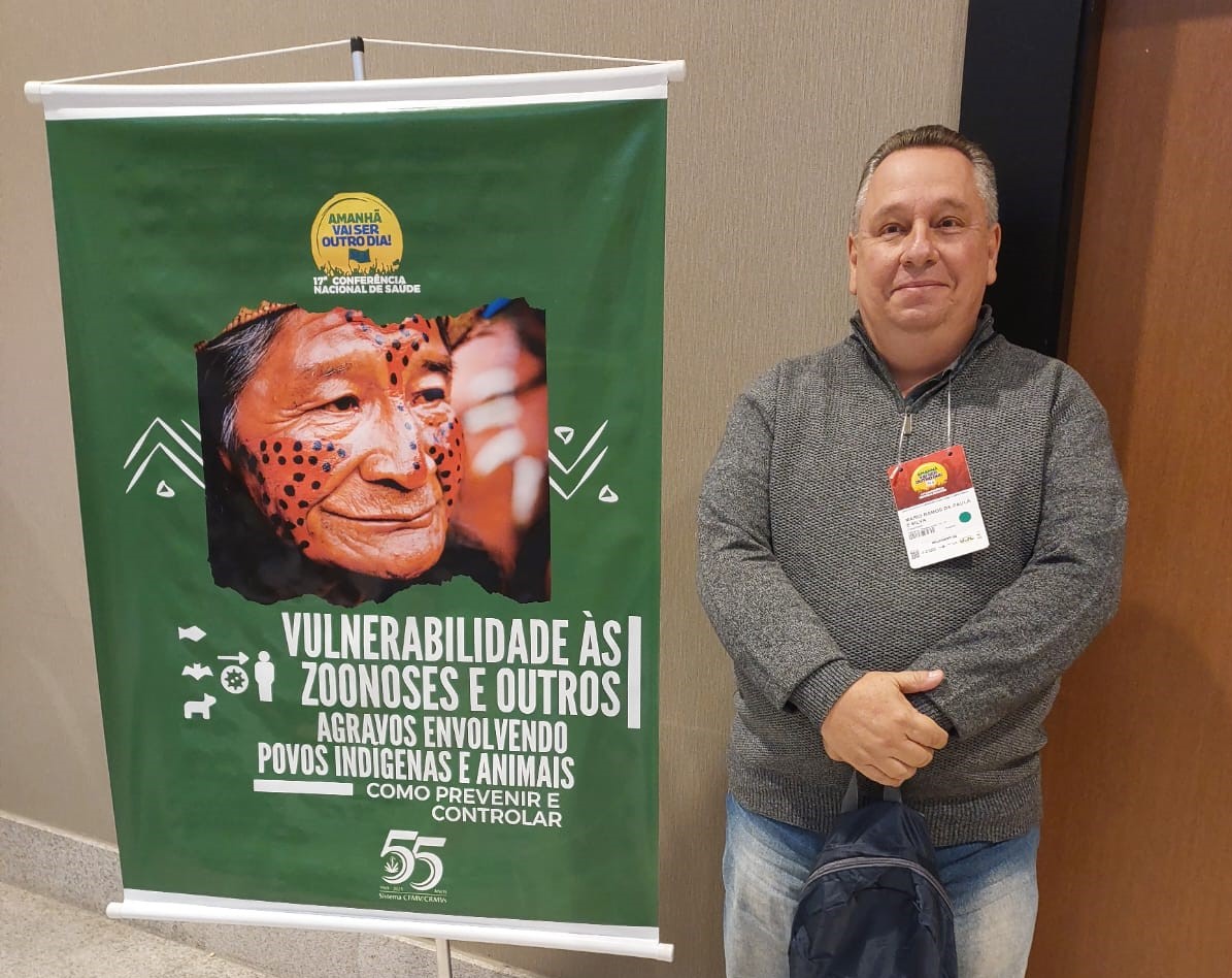 Na imagem aparece o integrante da Comissão Técnica de Saúde Pública do CRMV-SP. Ele é um homem de meia idade, com cabelos curtos e grisalhos. Ao lado dele há um banner verde sobre zoonoses e saúde indígena. No material há a imagem de um indígena com o rosto pintado tradicionalmente.