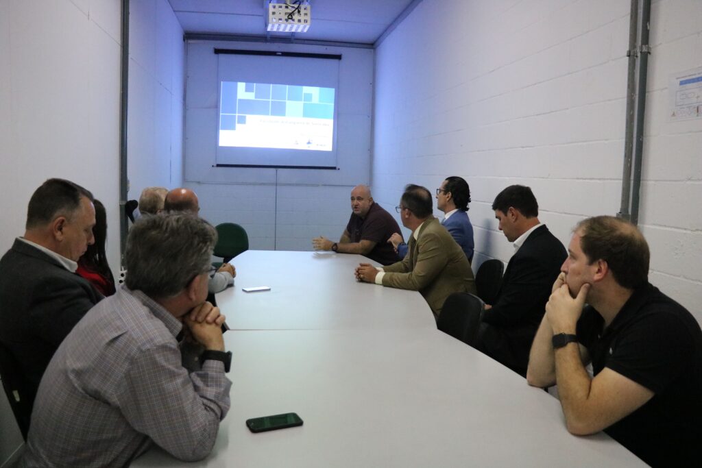 Na foto aparece a comitiva do CRMV-SP em sala de reunião com o coordenador do curso de Medicina Veterinária da Anhanguera. Estão todos sentados conversando ao entorno de uma mesa cinza. Ao fundo, há a projeção de um slide sobre o curso.
