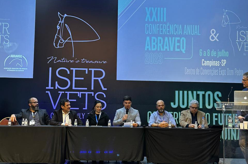 Os participantes da Conferência Anual da Associação Brasileira de Veterinários de Equídeos estão no palco, sentados, e o representante do CRMV-SP, Paulo Corte Neto, está falando ao microfone.