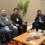 Na foto há cinco pessoas, sendo quatro homens e uma mulher, posando para a foto. Estão sentados, em volta de uma mesa de centro, em poltronas e sofá.