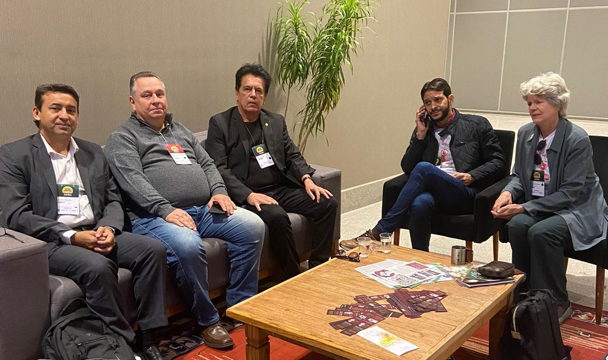 Na foto há cinco pessoas, sendo quatro homens e uma mulher, posando para a foto. Estão sentados, em volta de uma mesa de centro, em poltronas e sofá.