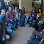 Na foto há nove pessoas, sendo cinco homens e quatro mulheres. Estão sentados em semicírculo posando para a foto.