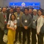 Na foto há oito pessoas, sendo cinco homens e três mulheres. Estão em pé, abraçados, posando para foto.