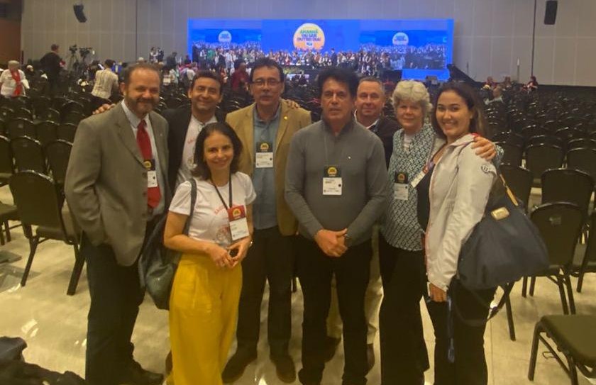 Na foto há oito pessoas, sendo cinco homens e três mulheres. Estão em pé, abraçados, posando para foto.