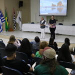 Na imagem aparecem a coordenadora do curso de graduação em Medicina Veterinária da Unesp e a representante regional do CRMV-SP. Elas estão em um palco de um auditório lotado de alunos. É possível ver a plateia sentada e de costas para a câmera. No palco há ainda um telão com projeção e as bandeiras do estado de São Paulo, do Brasil e do município de Araçatuba.