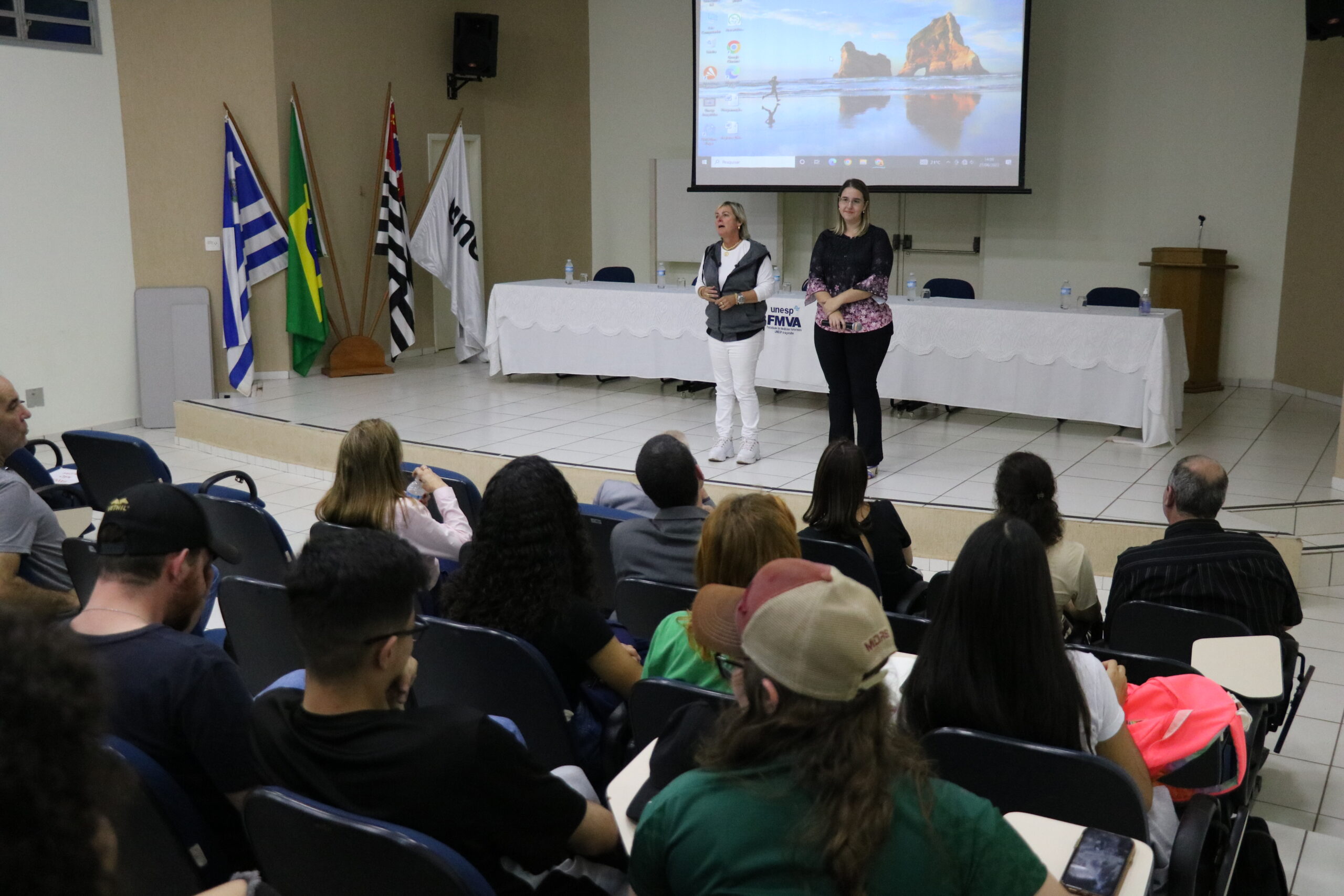 Na imagem aparecem a coordenadora do curso de graduação em Medicina Veterinária da Unesp e a representante regional do CRMV-SP. Elas estão em um palco de um auditório lotado de alunos. É possível ver a plateia sentada e de costas para a câmera. No palco há ainda um telão com projeção e as bandeiras do estado de São Paulo, do Brasil e do município de Araçatuba.