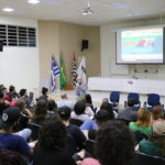 Na imagem aparece o auditório lotado de alunos. Estão todos sentados e voltados de costas para a câmera. A frente é possível ver o presidente do CRMV-SP ao microfone. Atrás dele, um palco, com mesa, bandeiras do Estado, do Brasil e do município de Araçatuba, além de um telão com projeto do site do CRMV-SP.