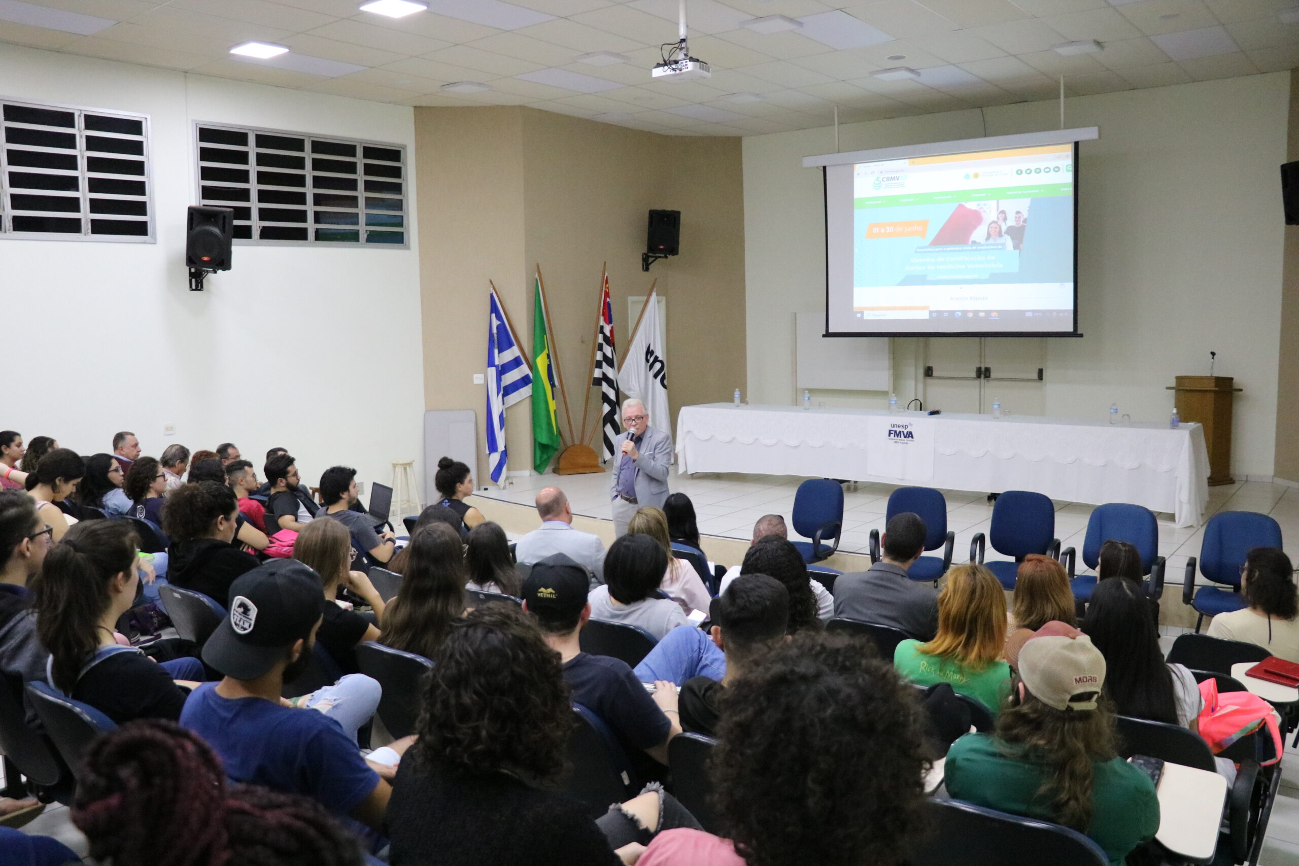 Na imagem aparece o auditório lotado de alunos. Estão todos sentados e voltados de costas para a câmera. A frente é possível ver o presidente do CRMV-SP ao microfone. Atrás dele, um palco, com mesa, bandeiras do Estado, do Brasil e do município de Araçatuba, além de um telão com projeto do site do CRMV-SP.