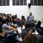 Na imagem aparece parte do auditório com alunos sentados nas carteiras. A sala é vista de perfil e no meio da plateia está o vice-presidente do CRMV-SP de pé e falando ao microfone.