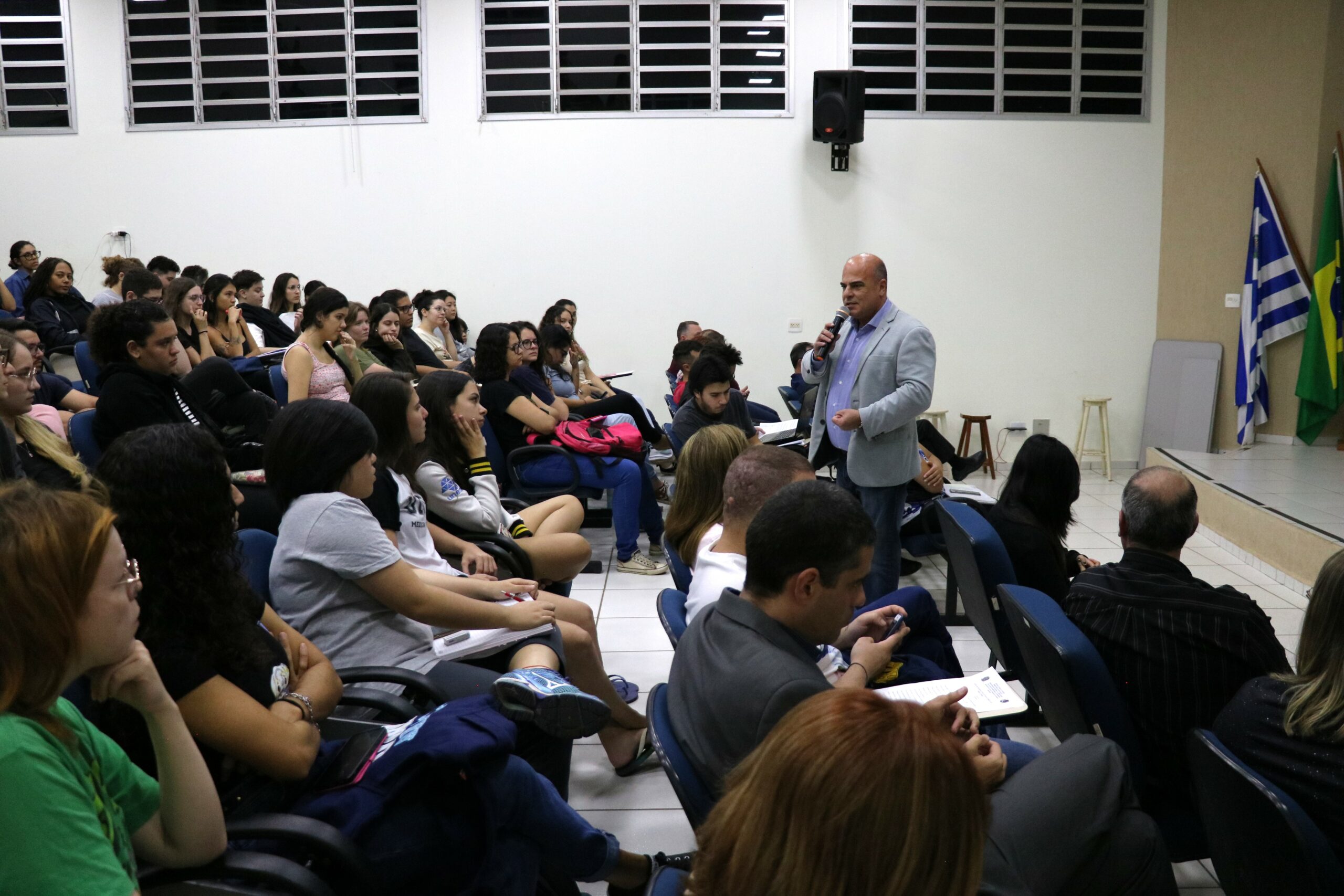 Na imagem aparece parte do auditório com alunos sentados nas carteiras. A sala é vista de perfil e no meio da plateia está o vice-presidente do CRMV-SP de pé e falando ao microfone.