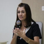 Na imagem aparece em destaque a tesoureira do CRMV-SP. Ela gesticula e fala ao microfone. Ela tem cabelos castanhos alinhados na altura do ombro e veste um macacão de cor preta e detalhe da gola em branco.