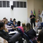 Na imagem aparece um auditório lotado de estudantes. A frente está o representante da Feveresp de pé ao microfone. Ele está de roupa preta e se se dirige ao presidente do Conselho, que está sentado também em frente a plateia.