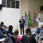 Na imagem aparece as primeiras fileiras do auditório. Os participantes aparecem de costas para a câmera. A frente está de pé o presidente da Comissão de Representantes Regionais do CRMV-SP. Ele veste um terno azul escuro e camisa cinza, usa óculos, e fala ao microfone. No canto inferior direito da foto aparece sentado em cadeiras azuis também o presidente do CRMV-SP. Ao fundo, as bandeiras de Araçatuba, Brasil, e São Paulo, assim como da própria Unesp.