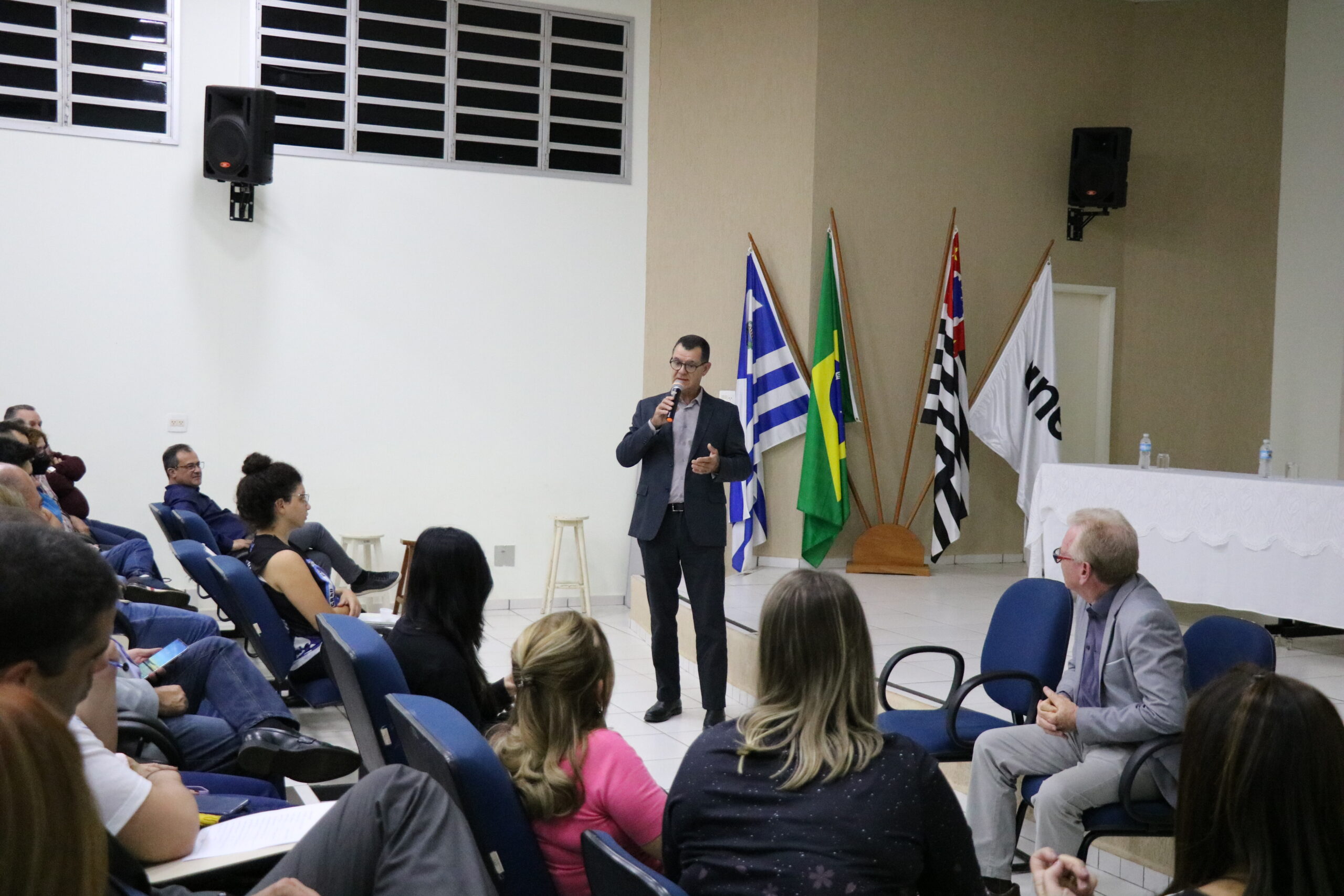 Na imagem aparece as primeiras fileiras do auditório. Os participantes aparecem de costas para a câmera. A frente está de pé o presidente da Comissão de Representantes Regionais do CRMV-SP. Ele veste um terno azul escuro e camisa cinza, usa óculos, e fala ao microfone. No canto inferior direito da foto aparece sentado em cadeiras azuis também o presidente do CRMV-SP. Ao fundo, as bandeiras de Araçatuba, Brasil, e São Paulo, assim como da própria Unesp.